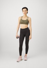 Sujetador deportivo verde oliva con logo negro, combinado con leggings negros de tiro alto. Zapatillas deportivas rosa claro, sosteniendo una botella de agua en la mano.