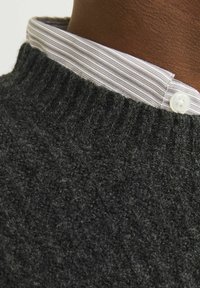 Pulover tricotat de culoare gri închis, cu guler ribat, purtat peste o cămașă cu guler în dungi albe și lila deschis, având un sistem de închidere cu nasturi.