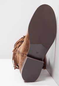 Botín de ante marrón con detalles de flecos, cremallera lateral y suela plana de goma. Talla marcada como 37 en la parte inferior de la suela. Parte superior texturizada.