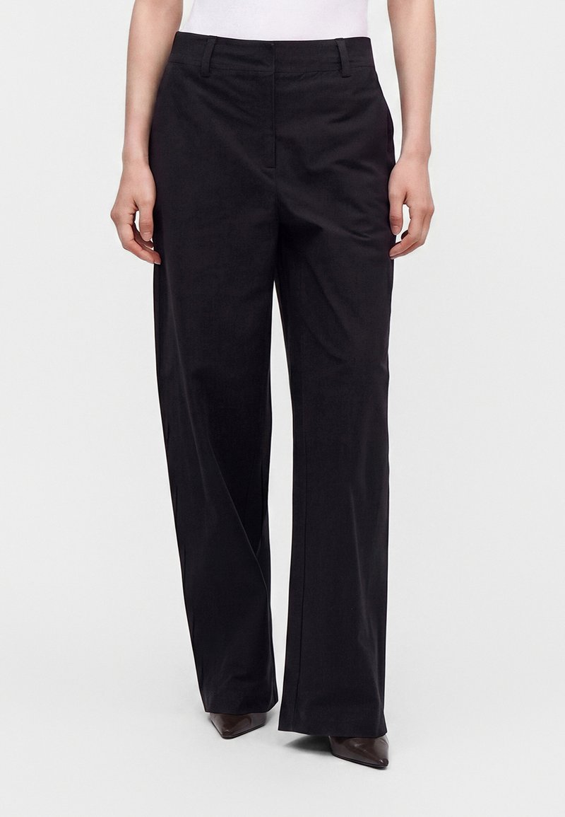 Personne portant un pantalon noir à jambes larges et des chaussures marron pointues, debout devant un fond blanc uni.