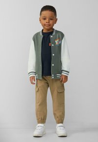 Groen en witte varsityjack met drukknoopsluiting, marineblauw T-shirt, khaki cargobroek en witte sneakers. Bevat geborduurd ontwerp.