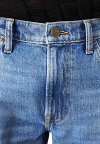 Jeans in denim azzurro chiaro con un taglio classico, caratterizzati da cuciture dorate, un bottone metallico con il logo "Lee" e design a cinque tasche.