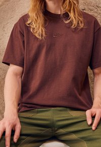 Hnědé bavlněné tričko s kulatým výstřihem a výšivkou loga Nike na hrudi, spárované se zelenými kalhotami se bočními kapsami.