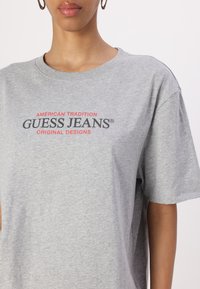 Šedé bavlněné tričko má kulatý výstřih, krátké rukávy a červený grafický text s nápisem "AMERICKÁ TRADICE ORIGINÁLNÍ DESIGNY GUESS JEANS."
