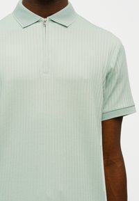 Polo en maille à manches courtes vert clair avec côtes verticales et col à fermeture éclair partielle porté par une personne à la peau foncée.