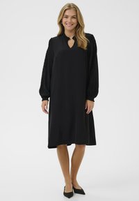 Vestido negro de mangas largas, con escote en forma de llave y acentos de encaje texturizado en las mangas. La tela suave cae hasta la mitad de la pantorrilla. Se lleva con tacones negros.
