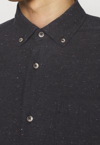 Mörk skjorta med knappar gjord av texturerat tyg med flerfärgade fläckar. Har en klassisk krage och grå knappar.