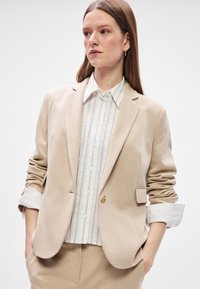 Bežový blejzer s textúrovanou tkaninou, s jedným gombíkom a klopným límcom. Nosený cez bielu košeľu s vertikálnym prúžkom a gombíkovým zapínaním.