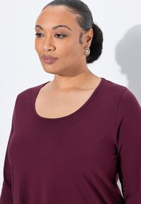 Burgunderfarbenes Langarmoberteil mit Rundhalsausschnitt, aus einem weichen Stoff gefertigt. Hat eine glatte Textur und eine taillierte Silhouette. Goldene Creolen sind sichtbar.