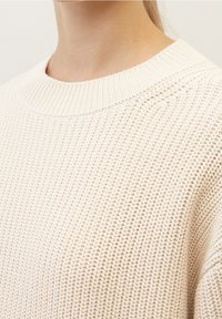 Maglione in maglia beige chiaro con texture a coste, ampia scollatura a girocollo e materiale morbido, che mostra una lavorazione intricata e un colore neutro.