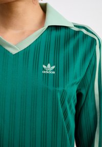Žalias dryžuotas ilgomis rankovėmis polo marškinėliai su plačia apykakle, ant krūtinės mažas baltas adidas logotipas ir kontrastingi šviesiai žali akcentai.