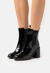Black patent leather ankle boots with a square toe, elastic side panels, and a chunky heel, featuring a subtle logo detail on the side.