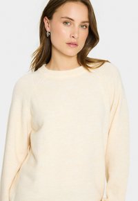 Kvinna med ljusbrunt hår som bär silverfärgade hoopörhängen och en krämfärgad tröja mot en enkel ljus bakgrund.