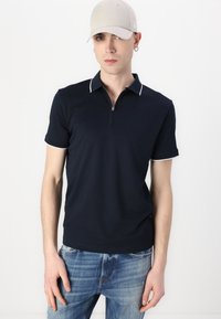 Tricou polo bleumarin cu guler cu fermoar de un sfert, având o margine albă pe guler și mansete, purtat cu blugi din denim ușor albastri.
