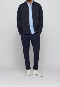 Man som bär en marinblå jacka och byxor, en ljusblå skjorta med knappar och vita högskurna sneakers, stående med händerna i fickorna.