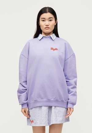 Jeune femme aux longs cheveux foncés portant un sweat-shirt lavande oversize par-dessus une robe à carreaux avec une broderie rouge à l’avant et à l’arrière.