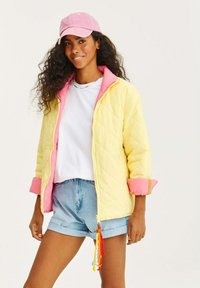 Prošívaná bunda v žluté barvě s růžovými akcenty, kombinovaná s bílým tričkem a modrými džínovými šortkami. Obsahuje barevné taháky na zip.