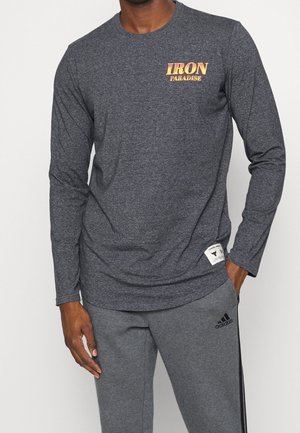 Mand iført en mørkegrå langærmet trøje med teksten "IRON PARADISE" og grå Adidas joggingbukser med sort logo og striber.