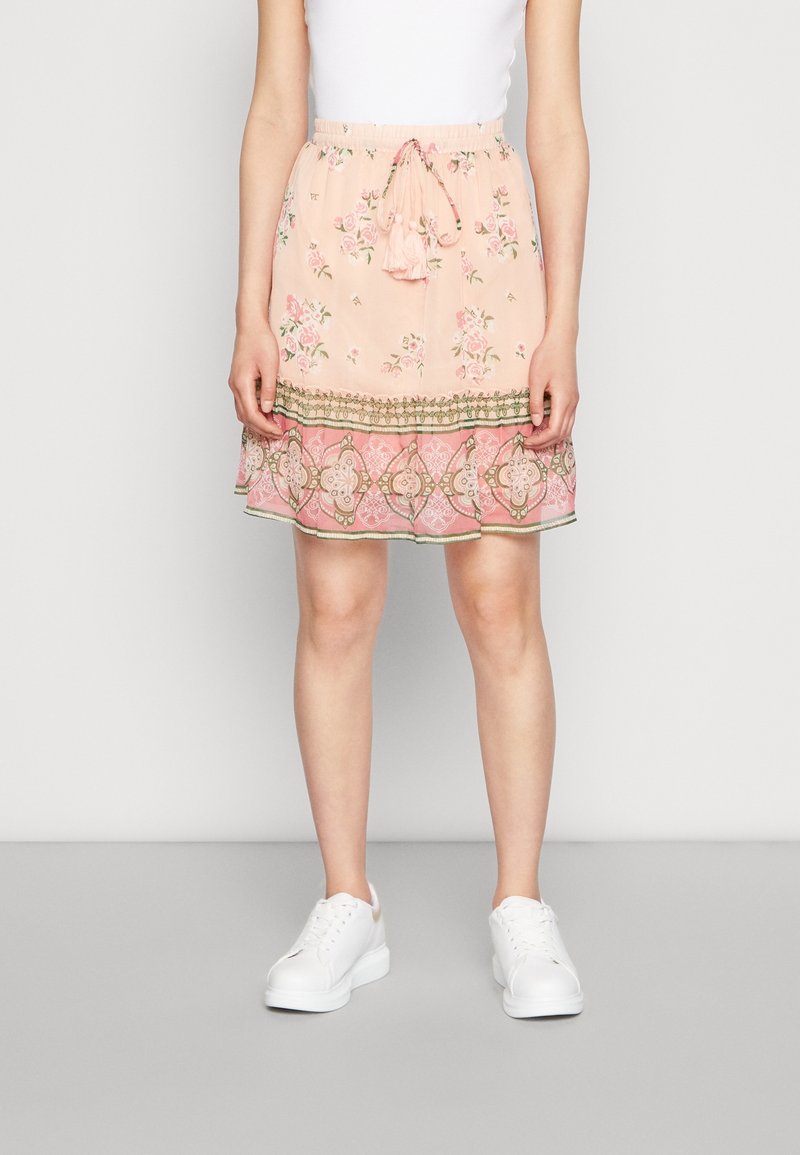 Femme portant une jupe florale rose clair avec une bordure décorative et des baskets blanches, debout devant un fond uni.