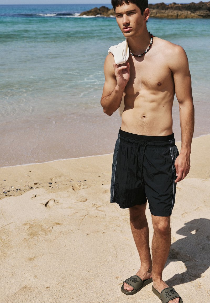Jeune homme torse nu portant un short de bain noir et des sandales, debout sur une plage de sable tenant une chemise blanche sur l'épaule près des vagues de l'océan.