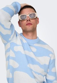 Lyseblå og hvid mønstret sweater med et skydesign, parret med beige solbriller med firkantede linser og tykke stel.