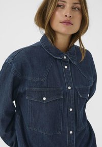 Donkerdenim overhemd met een puntige kraag, twee voorzakken en knoopsluiting. Bevat lichte stiksels en heeft een relaxte pasvorm.