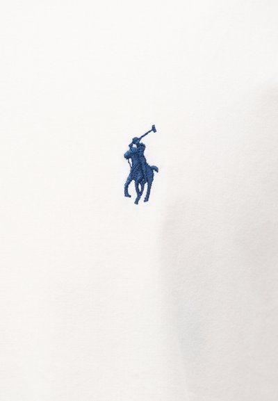 Modrá vyšívaná postava hráča póla na koni, ktorý drží zdvihnutý palicu, na bielom látkovom pozadí.