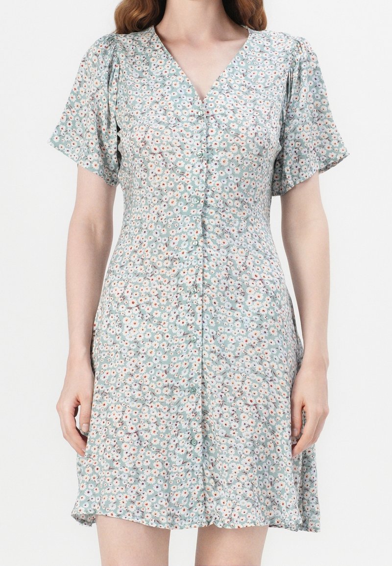 Femme portant une robe à manches courtes avec des boutons sur le devant, fond bleu clair et petit motif floral blanc et rouge.