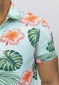 Chemise à manches courtes en tissu léger, avec une base vert menthe ornée de fleurs d'hibiscus rouges et orange vives et de grandes feuilles vertes.