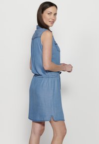 Abito in denim a maniche corte, di colore azzurro chiaro, con vita cintrata e spacco laterale. Presenta un dettaglio della tasca sul petto e una trama morbida.