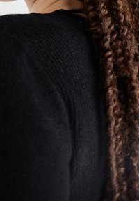 Pull en maille noir texturé avec un col rond, montrant un gros plan de l'épaule et du côté. Des cheveux noirs et ondulés tombent à côté.