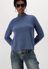 Blue mock neck sweater in knit fabric with long sleeves, paired with faded denim jeans. Features a relaxed silhouette and soft texture.