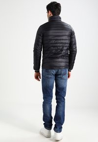 Svart puffjacka med horisontella sömmar, hög krage och fast textur, i kombination med blå jeans och vita sneakers, sedd bakifrån.