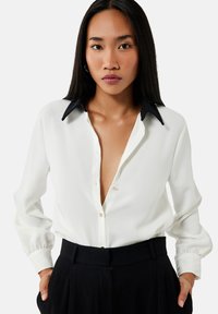 Blusa blanca con escote en V profundo y cuello negro, con mangas largas y cierre de botones, hecha de una tela ligera.