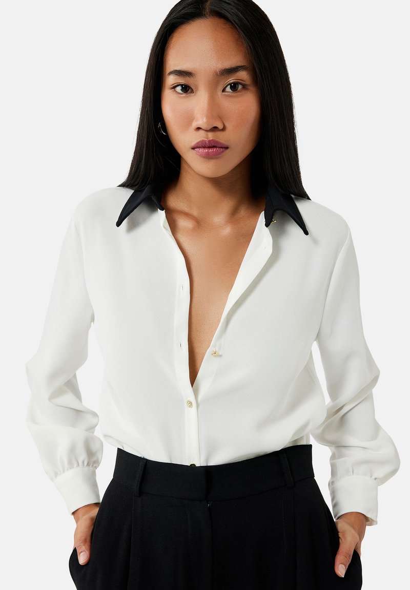 Blusa blanca con escote en V profundo y cuello negro, con mangas largas y cierre de botones, hecha de una tela ligera.