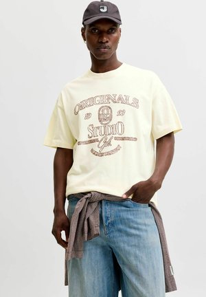 Joven vestido con una camiseta gráfica beige, vaqueros azul claro, una camisa de cuadros atada a la cintura y una gorra de béisbol negra.