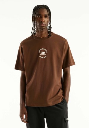 Giovane uomo con capelli intrecciati che indossa una maglietta marrone New Balance con logo bianco e pantaloni neri, in piedi davanti a uno sfondo bianco.