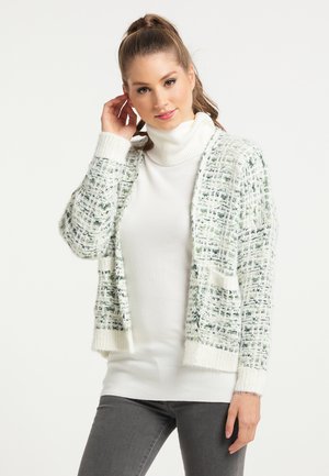 Vrouw draagt een witte coltrui en een gestructureerde gebreide cardigan met groen-wit patroon, poseert met hand bij het oor.