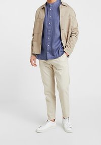 Chaqueta beige clara sobre una camisa azul de botones, combinada con pantalones color crema y zapatillas blancas; estilo casual, corte moderno y diseño simple.