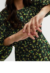 Zwarte jurk met geel bloemenpatroon en groene accenten. Lange mouwen met gathers bij de manchetten. Handen op de taille, met een roze manicure.
