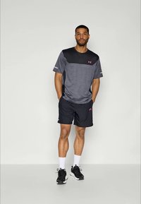 Hombre de pie con las manos en los bolsillos, lleva una camiseta atlética negra y gris, pantalones cortos negros, calcetines blancos y zapatillas de correr negras sobre un fondo llano.