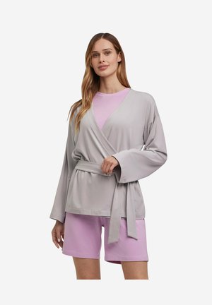 Donna che indossa una giacca avvolgente grigio chiaro con cintura sopra una maglia malva e pantaloncini abbinati malva, in piedi davanti a uno sfondo bianco neutro.