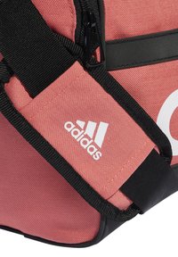 Borsa adidas rosa con spalline e zip nere, caratterizzata da un grande logo bianco e tessuto testurizzato. Vista laterale che mostra cuciture dettagliate e accenti.