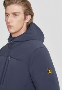 Veste à capuche navy avec une texture lisse, présentant une poche poitrine zippée et un patch logo sur la manche. Design simple et moderne.