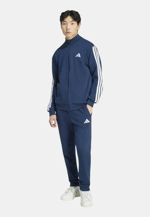Giovane uomo in piedi che indossa una tuta Adidas blu navy con strisce bianche e scarpe da ginnastica bianche su uno sfondo bianco uniforme.