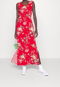 Žena v bezrukávových červených květovaných maxi šatech s bočním rozparkem, bílé vysoké tenisky a držící malou zelenou kabelku.