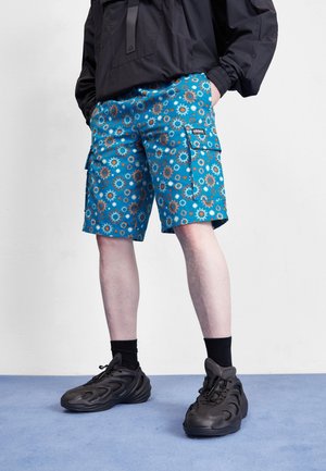 Person trägt blaue Cargo-Shorts mit Sonnenmustern, schwarze Sneakers, schwarze Socken und eine schwarze Jacke und steht auf einem blauen Boden.