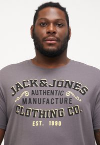 Pelēks kokvilnas T-krekls ar lieliem melniem un dzelteniem drukātiem burtiem, kas saka "JACK & JONES AUTHENTIC MANUFACTURE CLOTHING CO. EST. 1990."