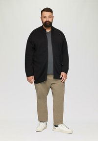 Schwarze Zip-Up-Strickjacke über einem grauen Shirt, kombiniert mit beigen Hosen und weißen Sneakers. Glattes Material, entspannte Passform, minimalistisches Design.