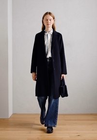 Manteau en laine noir avec des revers en pointe, porté sur une chemise rayée, associé à un jean évasé et des chaussures en cuir verni noir. Portant un sac texturé.
